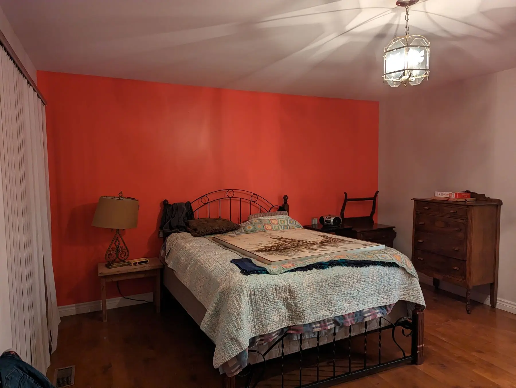 Before — A furnished bedroom with a bright orange accent wall, dark metal bed frame, and hanging light fixture.