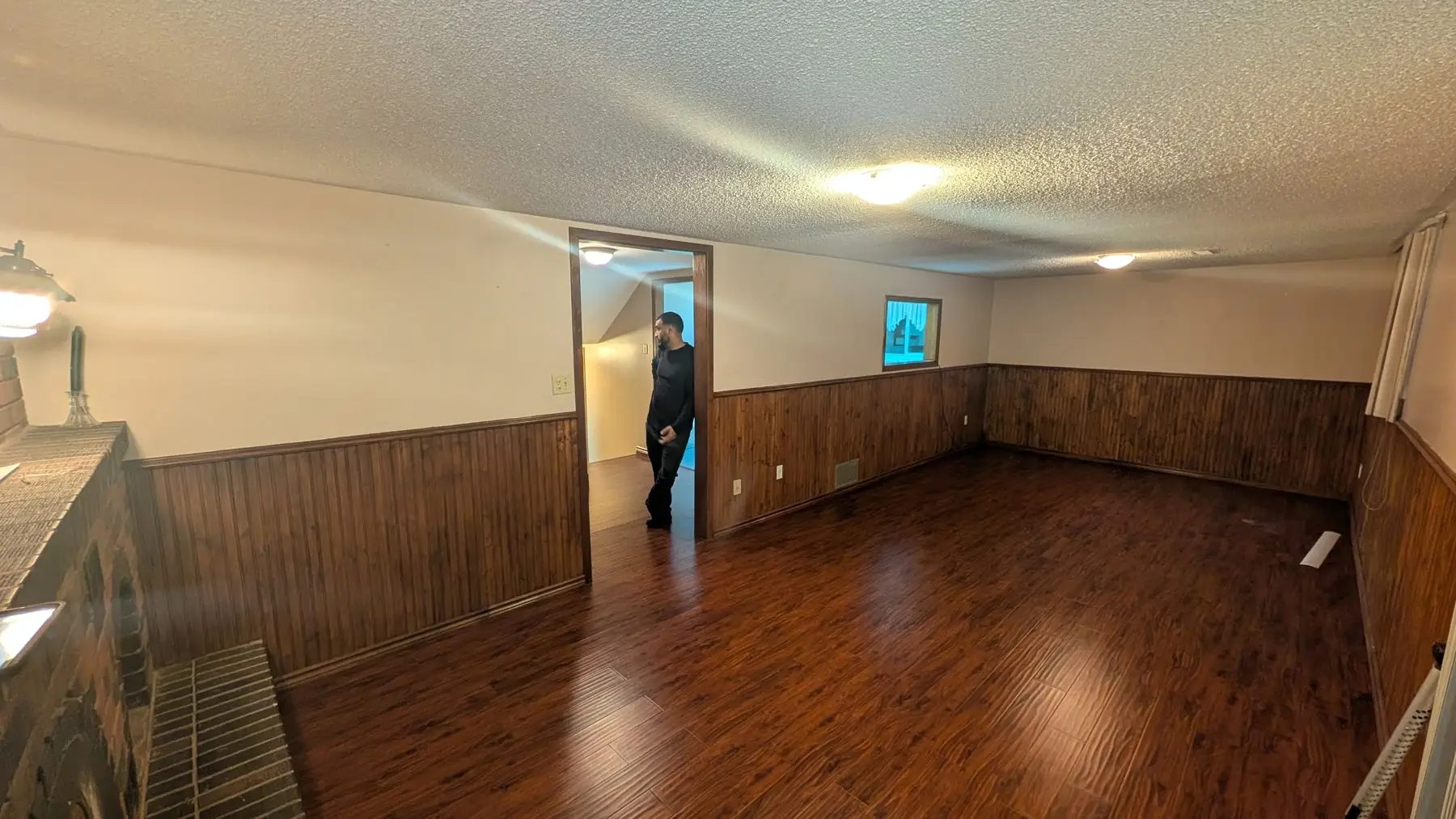 Before — Basement interior before renovation featuring wood wainscoting, brick fireplace, dark laminate flooring, and a worker standing in the doorway.