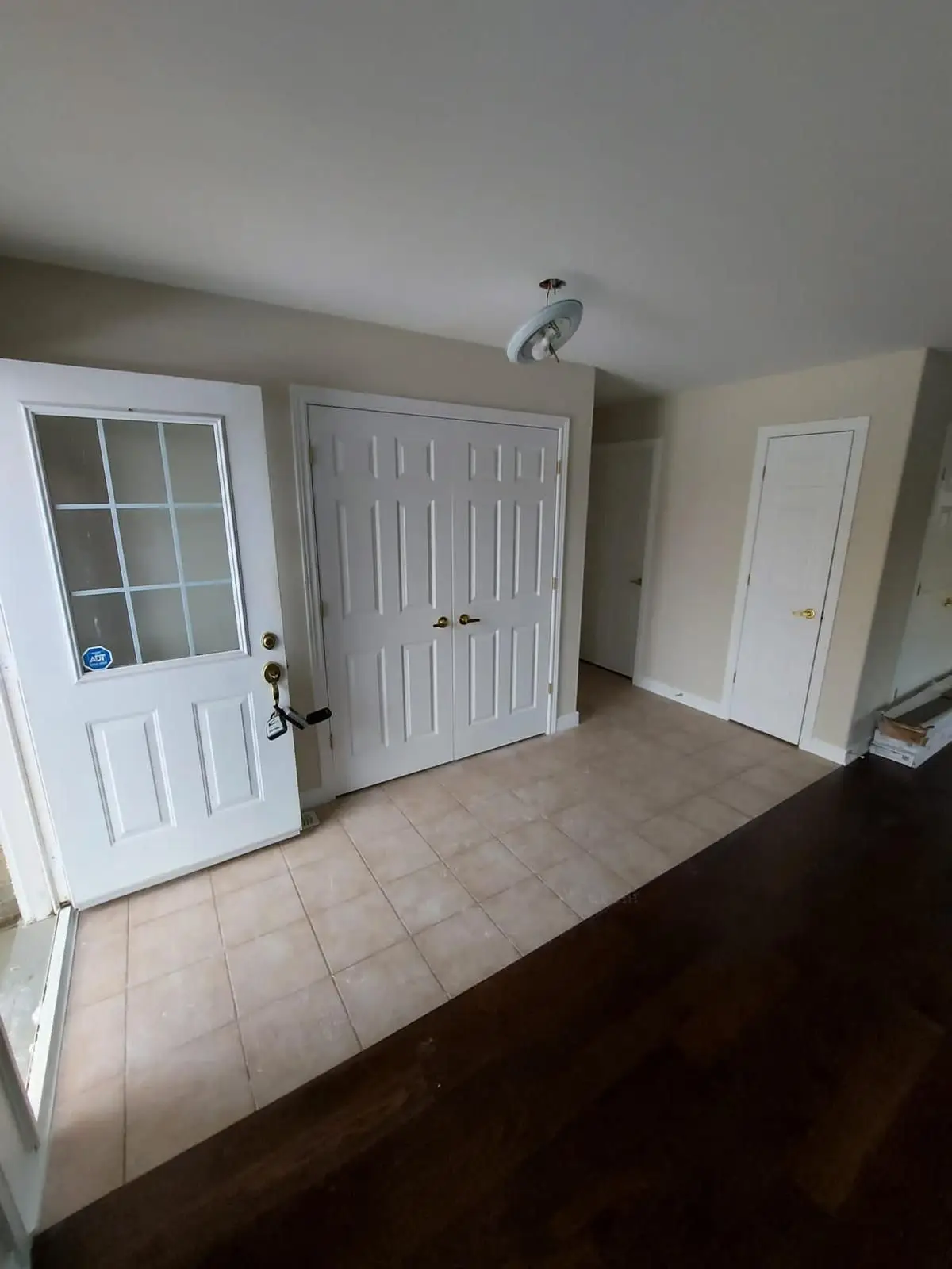 Before — Wide shot of an entryway with tiled floor, front door, closet doors, and transition to hardwood flooring.