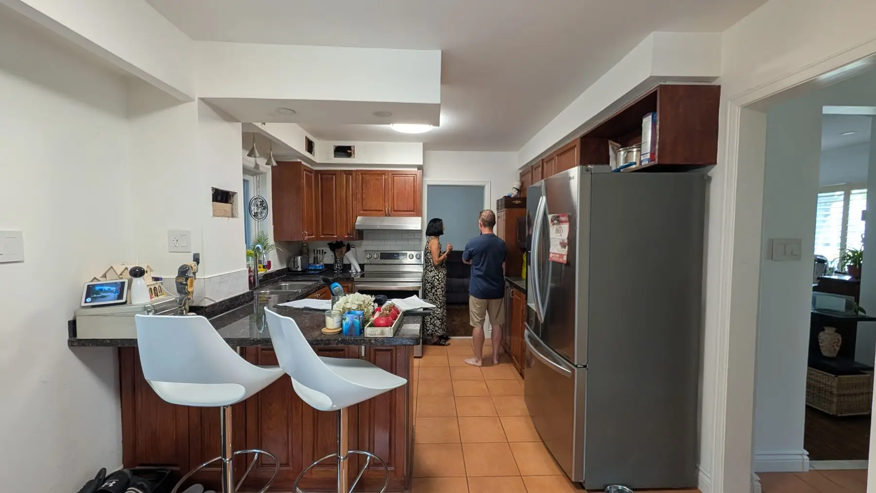 Before — Wide shot of existing kitchen with wooden cabinets, stainless steel appliances, and two people standing.