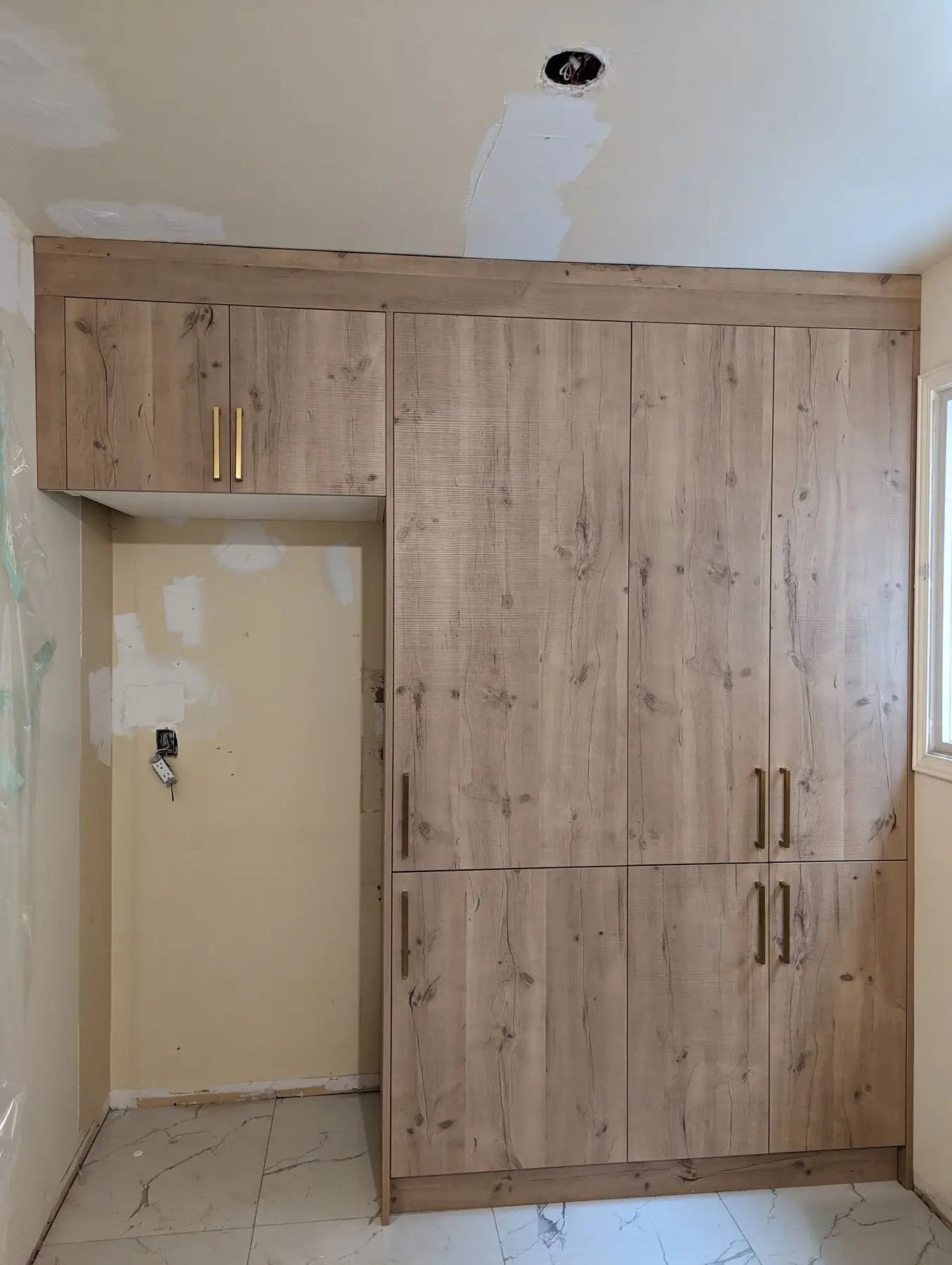 Finishing — Wood-grain kitchen cabinetry installed next to an unfinished appliance recess with drywall patching underway.