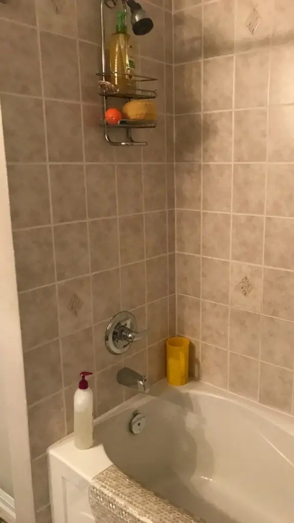 Before — A pre-construction view of a beige tiled bathtub and shower enclosure with a hanging caddy.