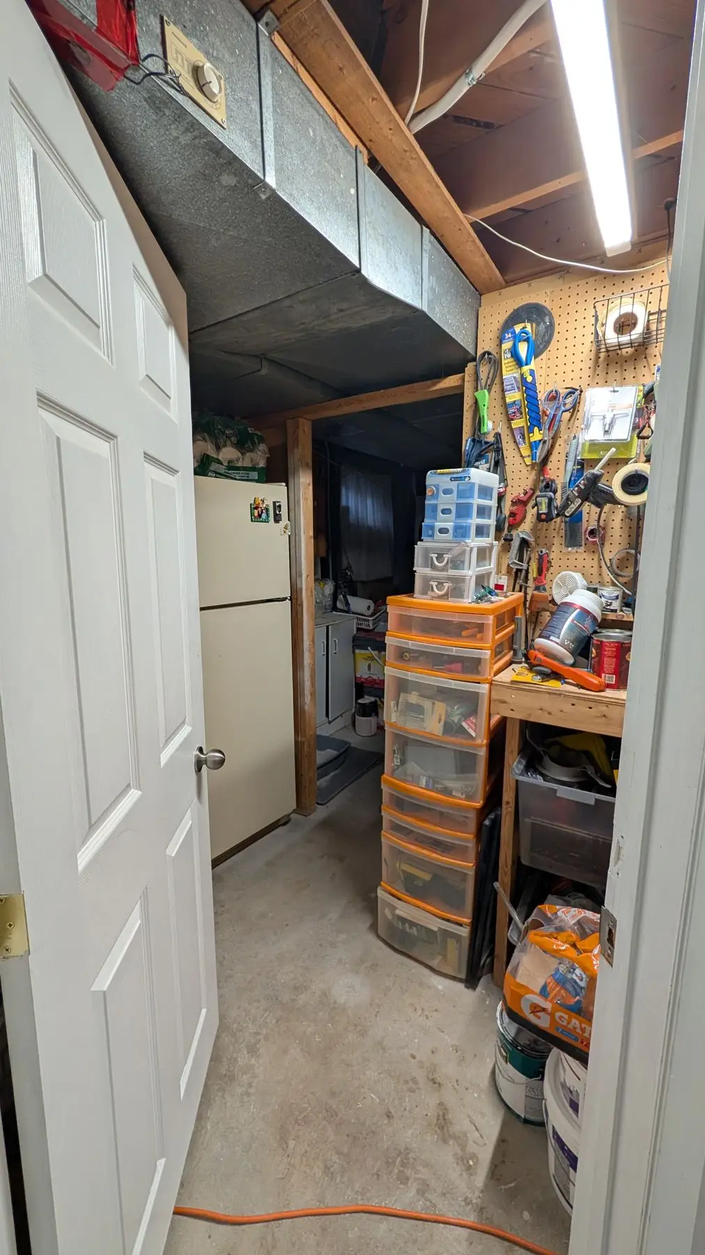 Before — Basement utility and storage area viewed through a doorway, featuring a pegboard wall and storage drawers.