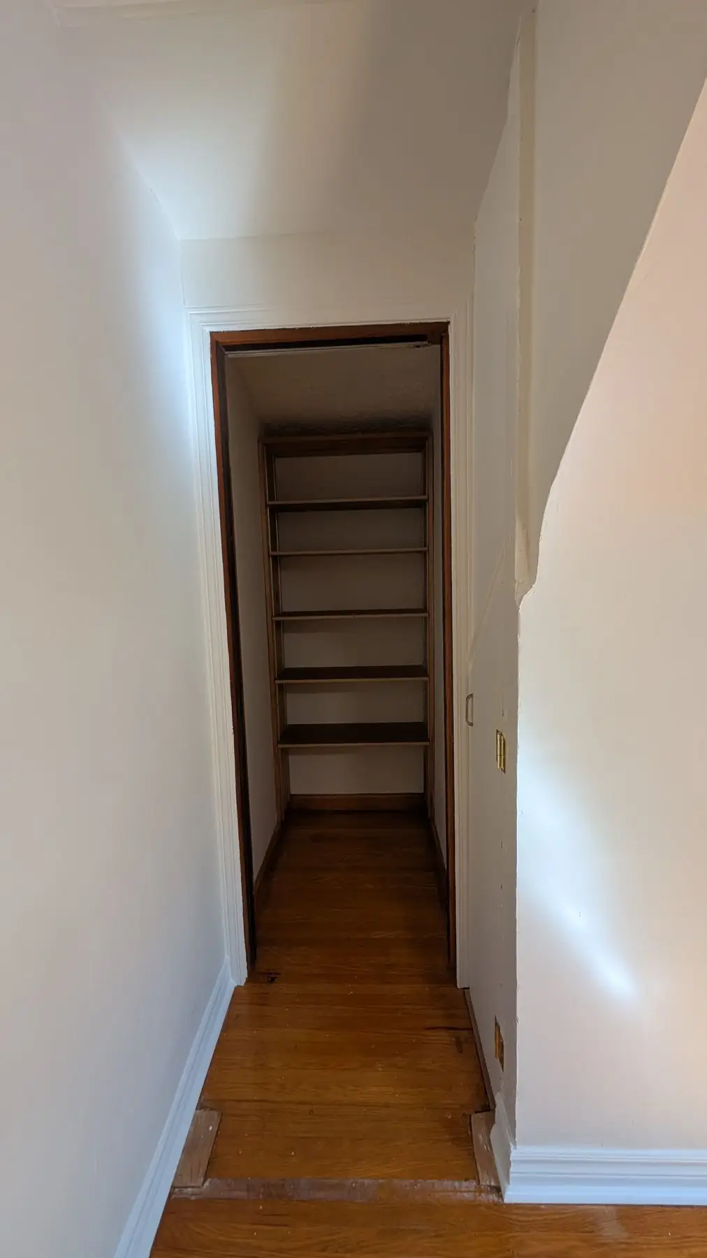 Before — View through a doorway into a small storage space with built-in wooden shelves and hardwood floors.