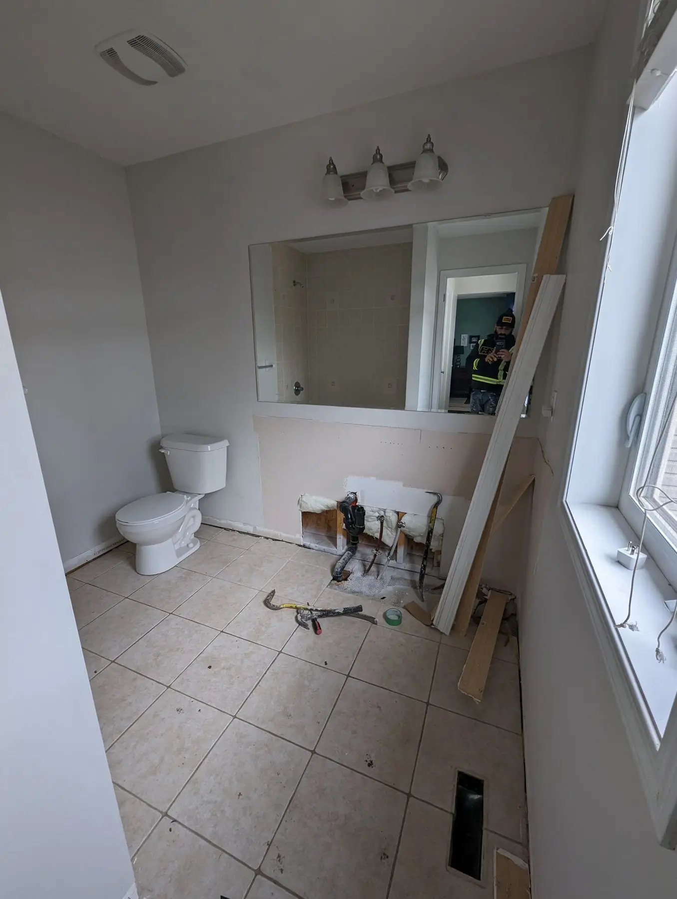 Rough-in — Bathroom during vanity removal with exposed plumbing, tools on the floor, and worker visible in mirror.