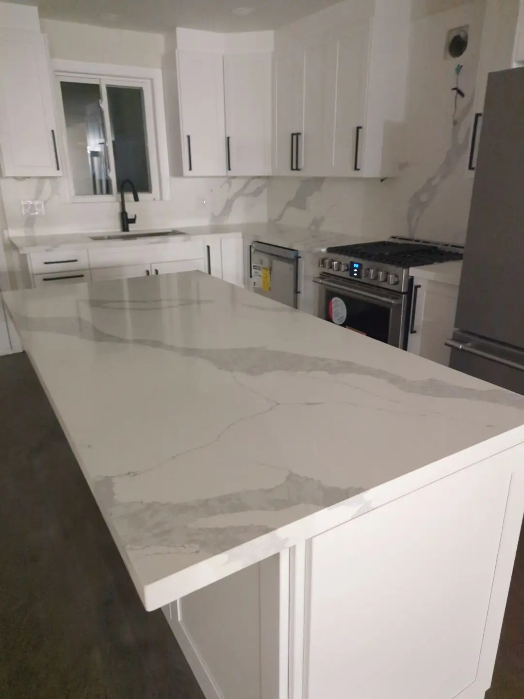 Finishing — Kitchen corner view showing installed white cabinetry, quartz island countertop, marble backsplash, and stainless steel appliances.