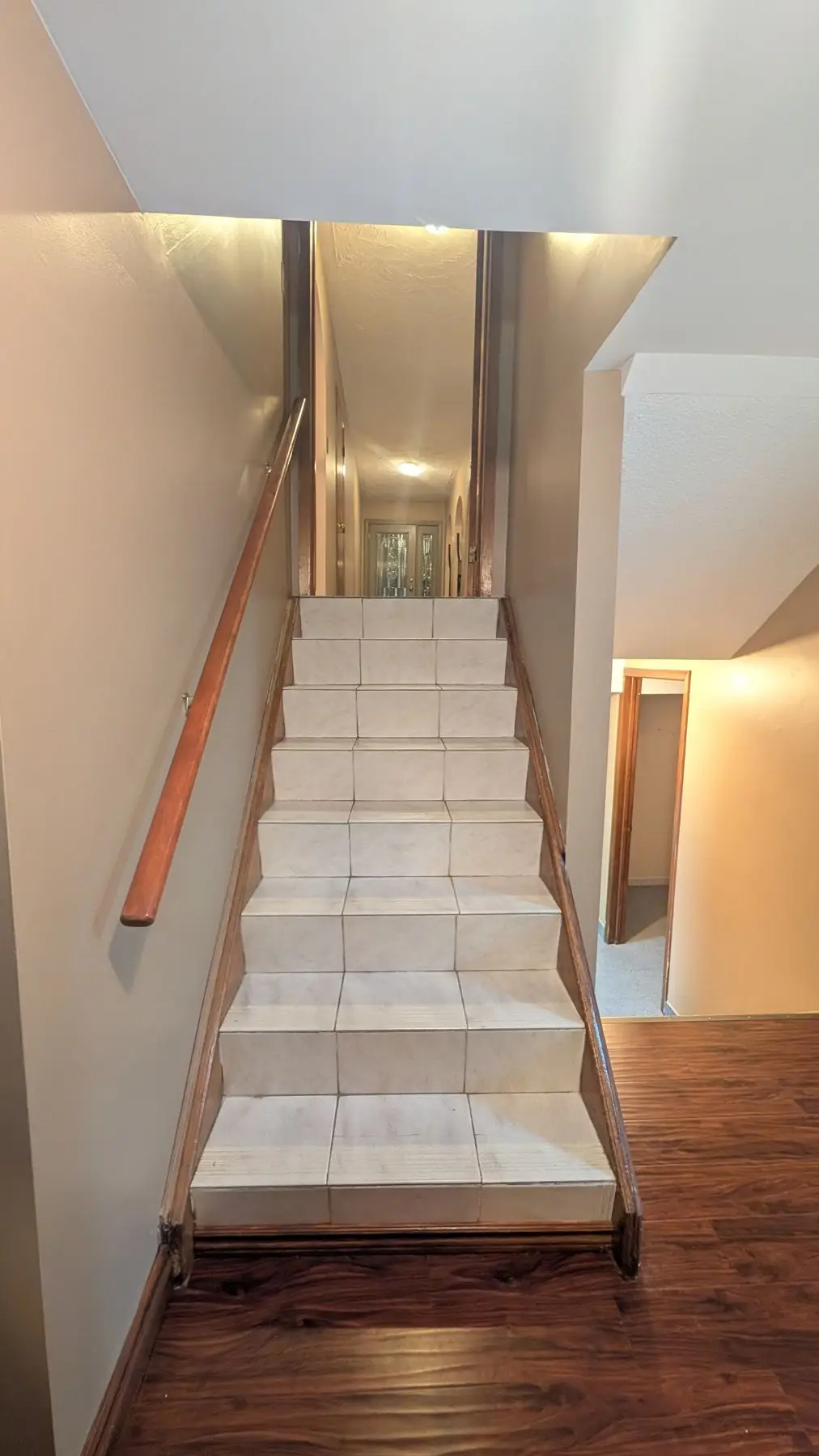 Finished — Straight flight of stairs finished with light ceramic tiles and a wooden handrail, leading up to a hallway.