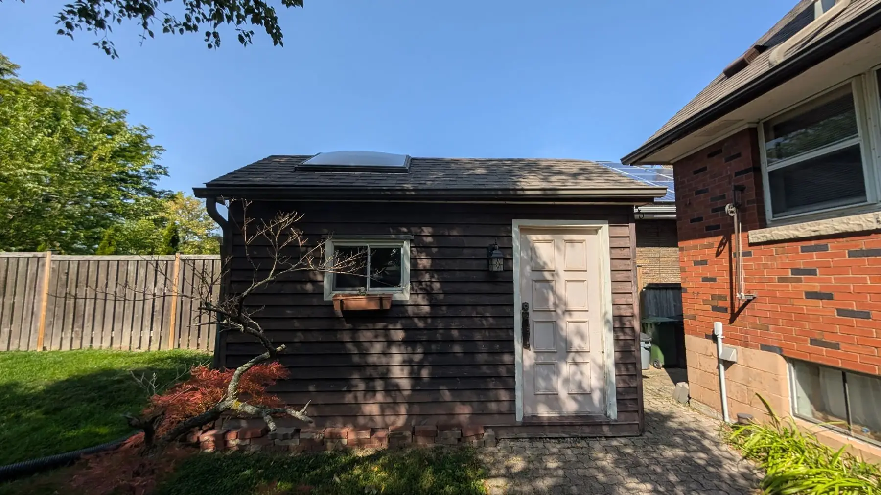 Finished — Exterior view showing a detached brown wooden shed with a skylight adjacent to a brick house.
