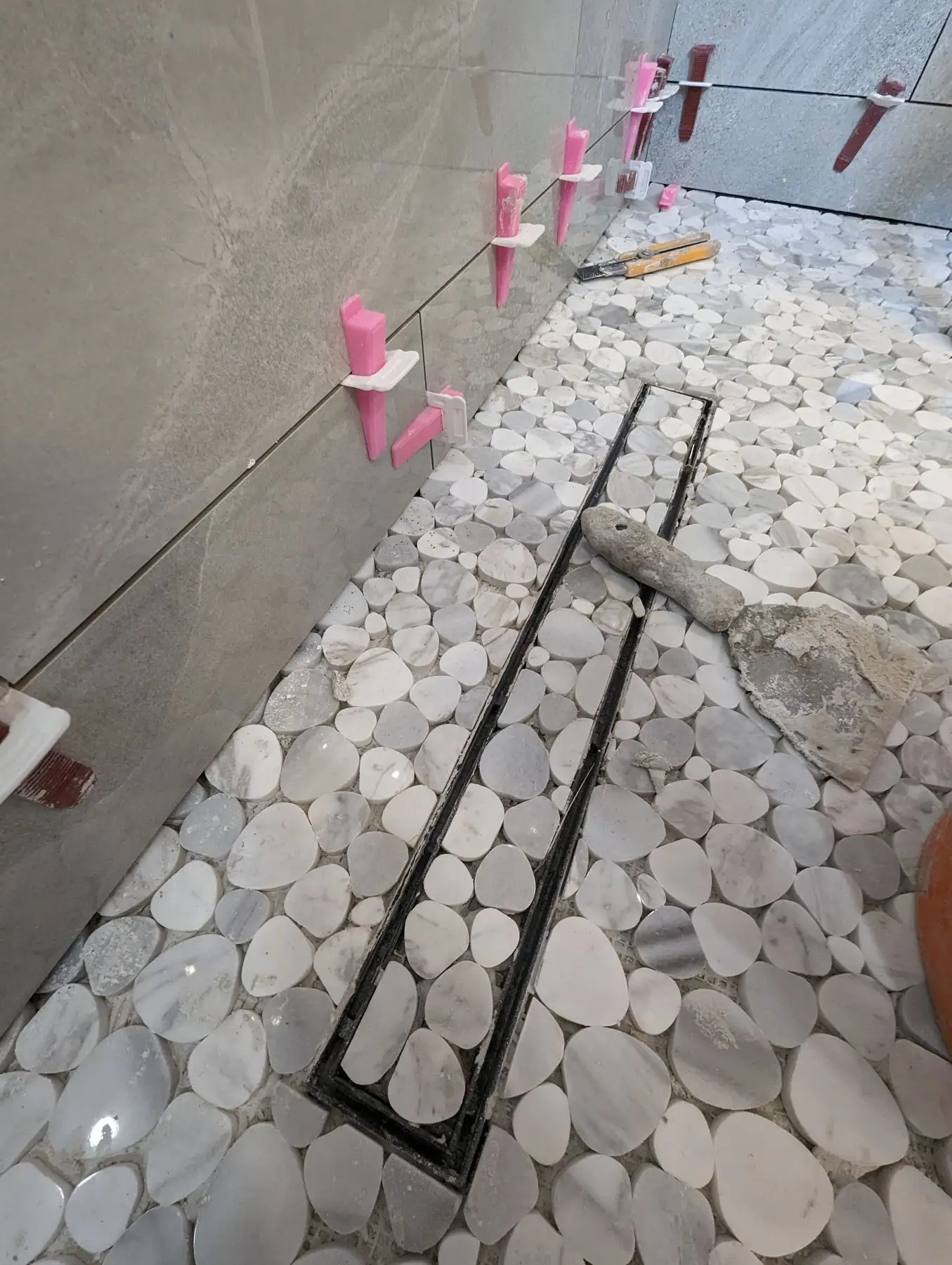 Finishing — Shower stall installation showing pebble tile floor, linear drain, and large format wall tiles with leveling spacers.