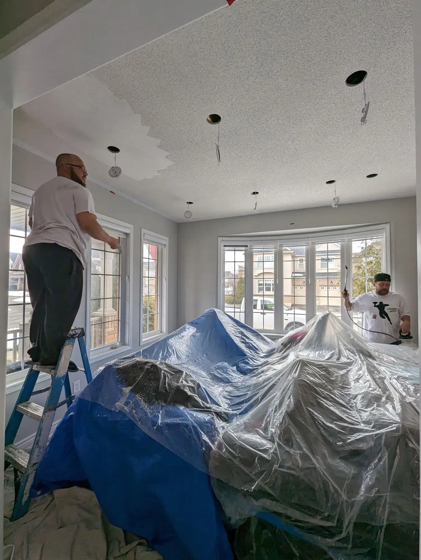 Insulation & drywall — Two workers painting and preparing the ceiling in a living room with draped furniture and a bay window.
