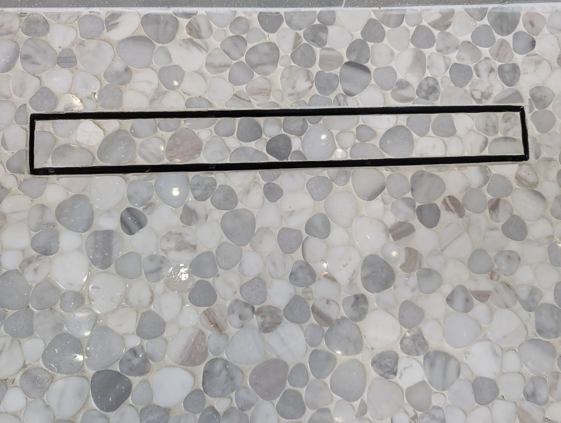 Finishing — Close-up of a shower floor featuring grey and white pebble mosaic tile and a black linear drain.