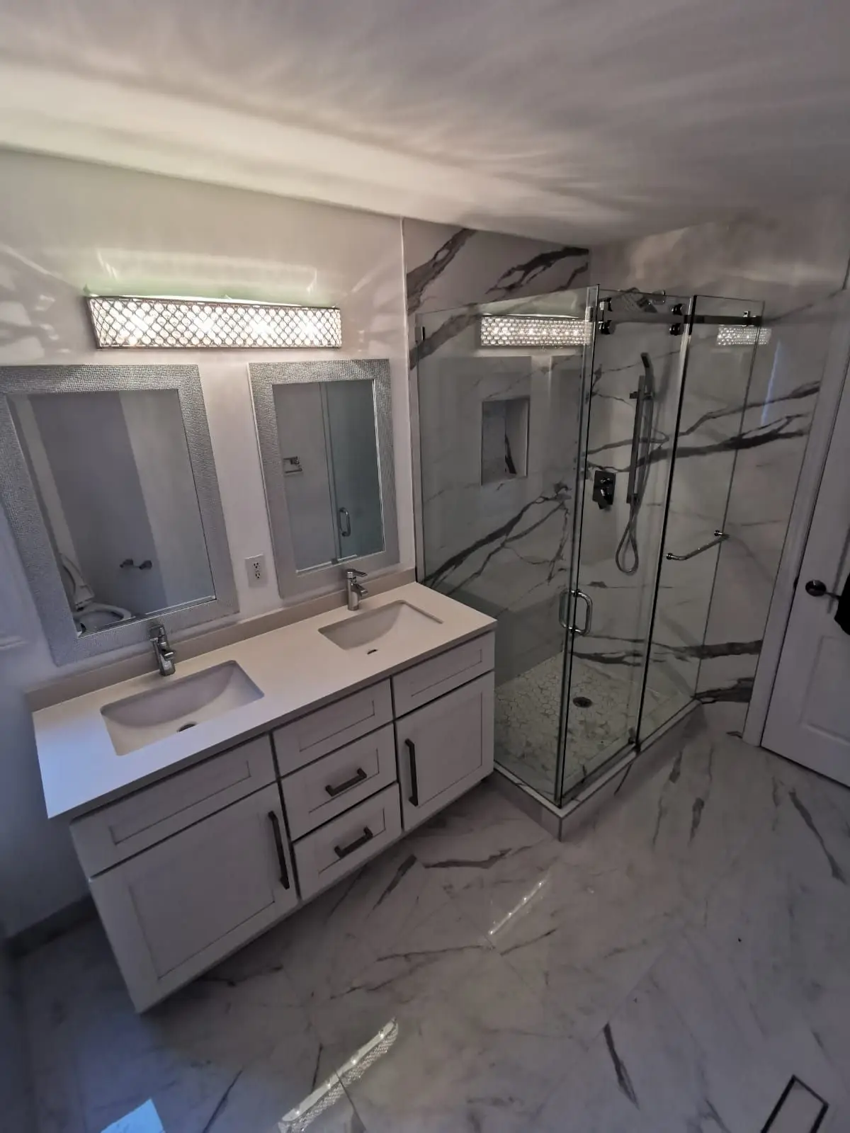 Finished — Completed modern bathroom featuring a double vanity, large glass shower, and marble-look tiles.