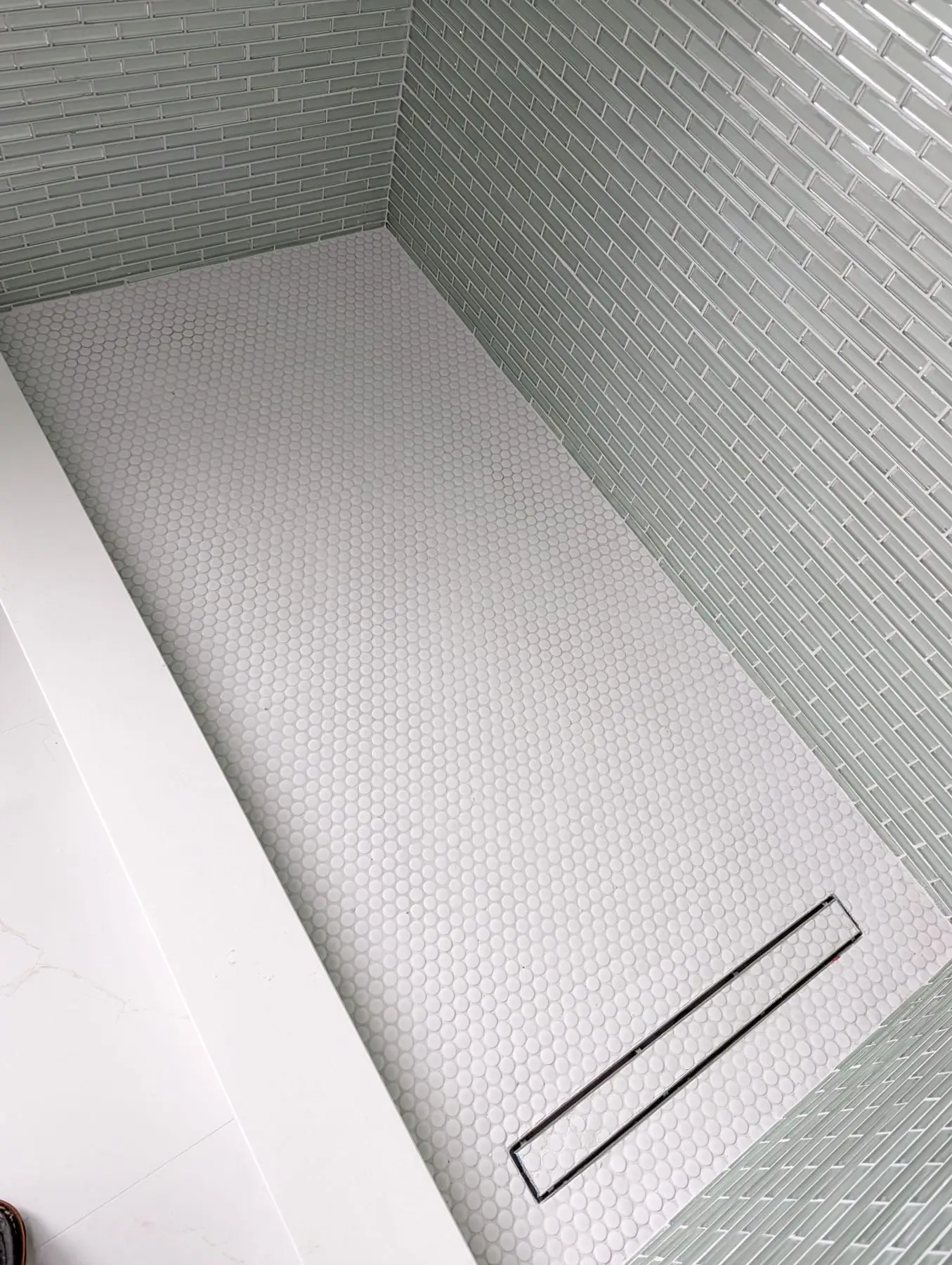 Finishing — High-angle shot of a newly tiled shower base featuring white penny tile and a linear drain.