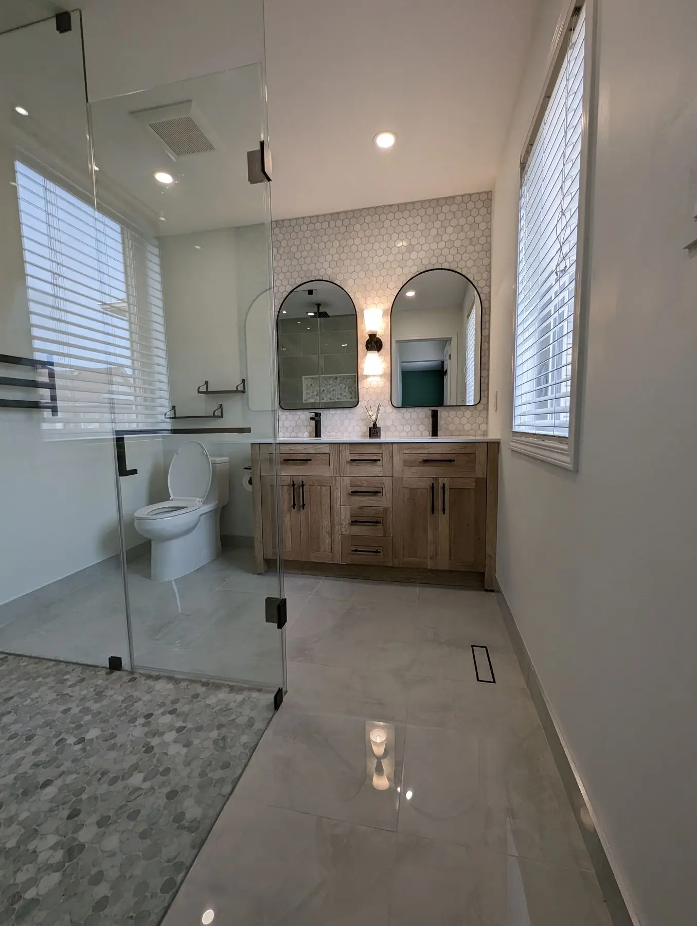 Finished — Completed modern bathroom featuring a double vanity with arch mirrors, glass walk-in shower, and hex tile backsplash.
