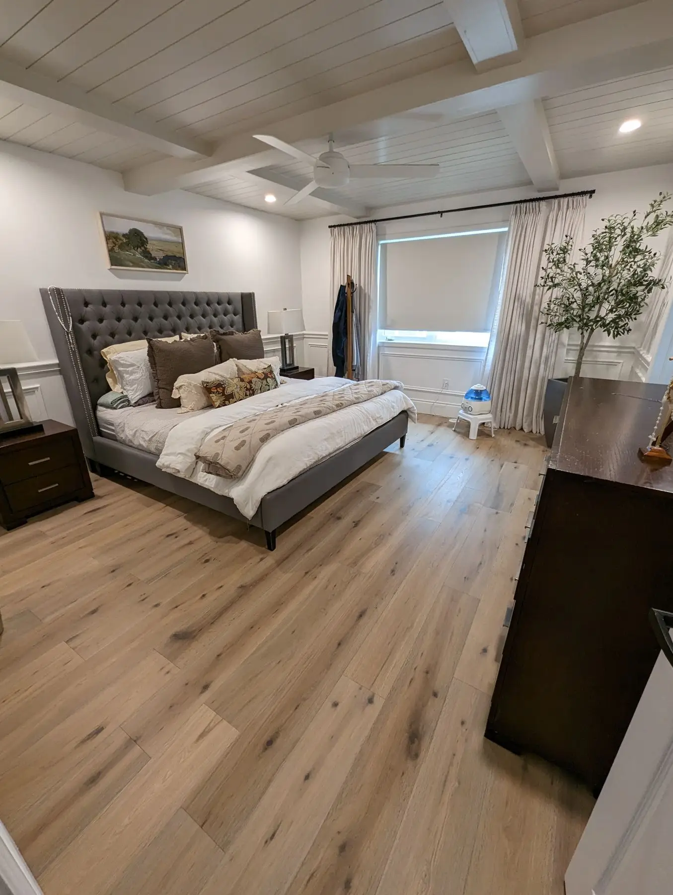 Finished — Wide corner shot of a completed bedroom with hardwood floors, a tufted bed, and ceiling beams.