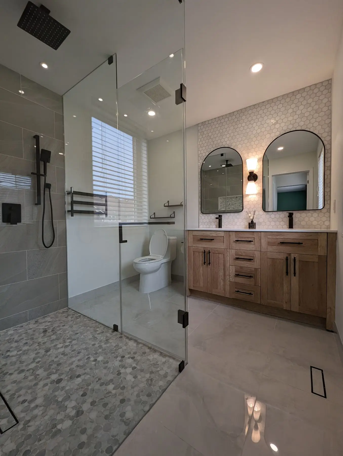 Finished — Completed modern bathroom featuring a double vanity with arched mirrors, large glass shower, and pebble tile flooring.