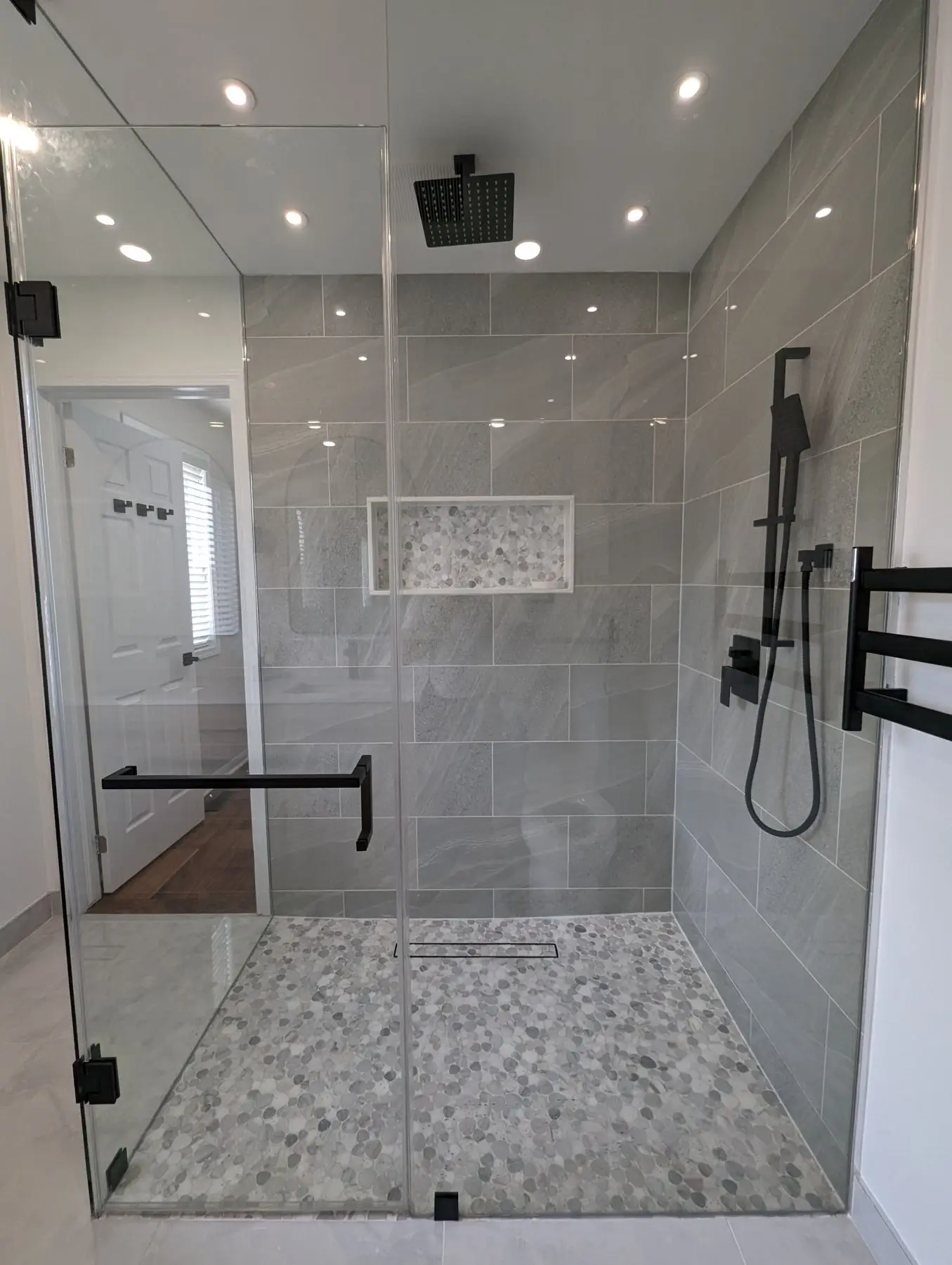 Finished — Completed modern walk-in shower with grey tiled walls, pebble floor, black fixtures, and glass enclosure.
