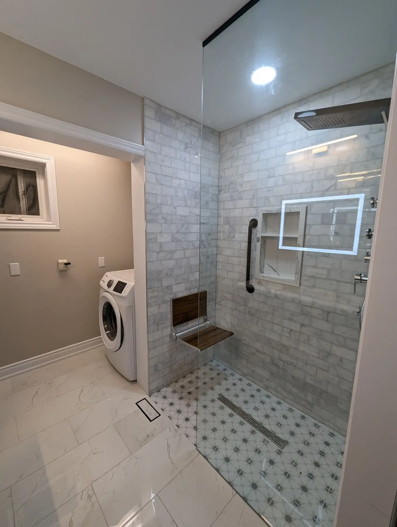 Finished — Completed modern bathroom with tiled walk-in shower, glass partition, wooden fold-down bench, and adjacent laundry room.