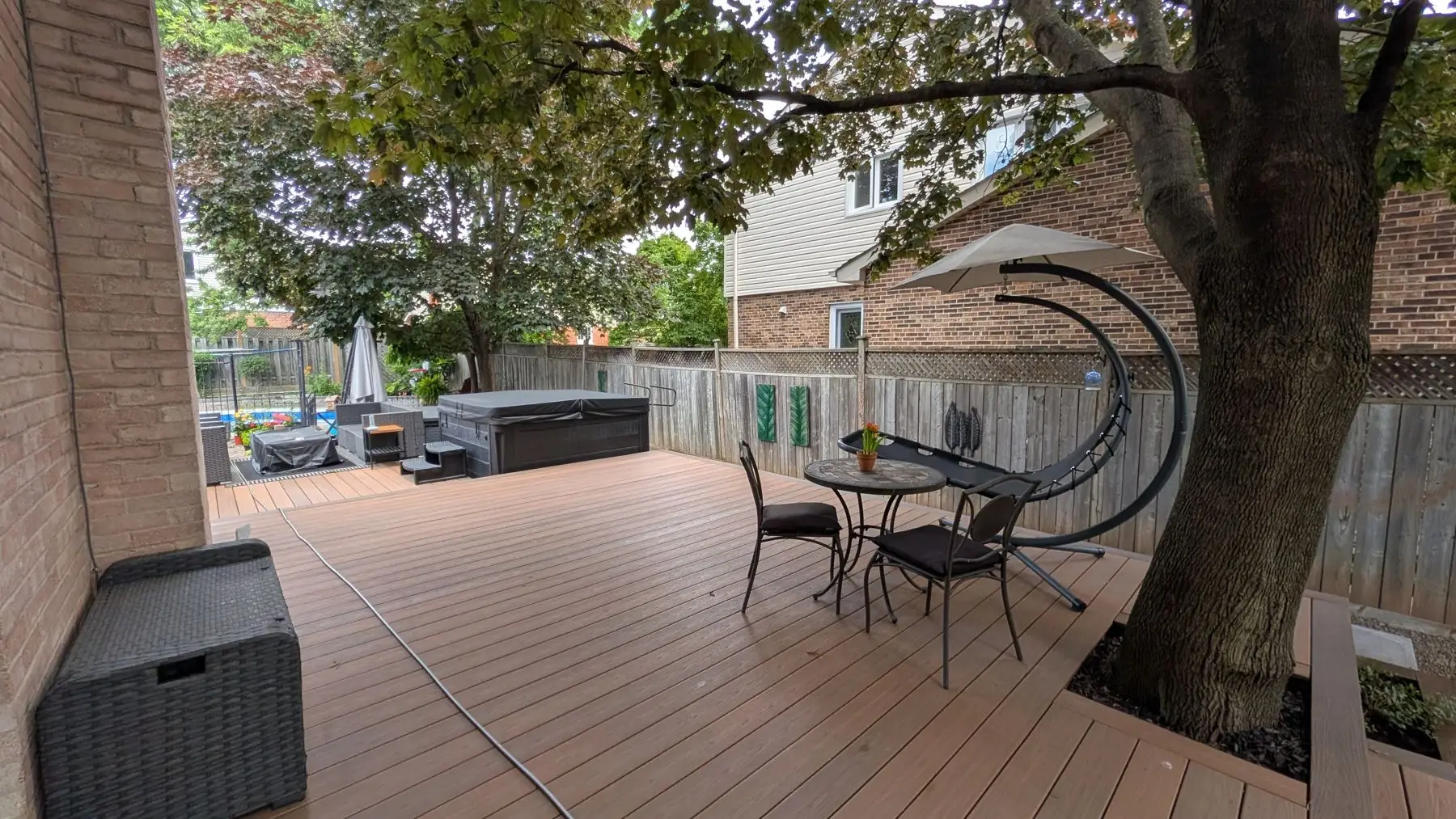 Finished — Completed multi-level wooden backyard deck featuring a hot tub, patio furniture, and a mature tree integrated into the design.